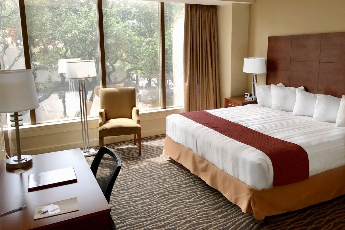A hotel room with a large bed, a desk with a lamp, a chair, and a view of trees through the window.