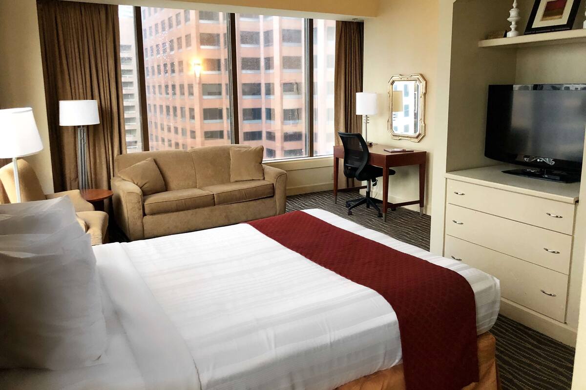 A hotel room with a large bed, a desk with a chair, a television, and a view of a building through the window.