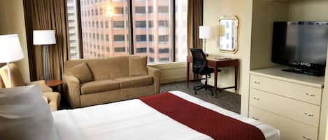 Habitación de hotel con una cama grande, un escritorio con una silla, televisión y vista a un edificio desde la ventana.