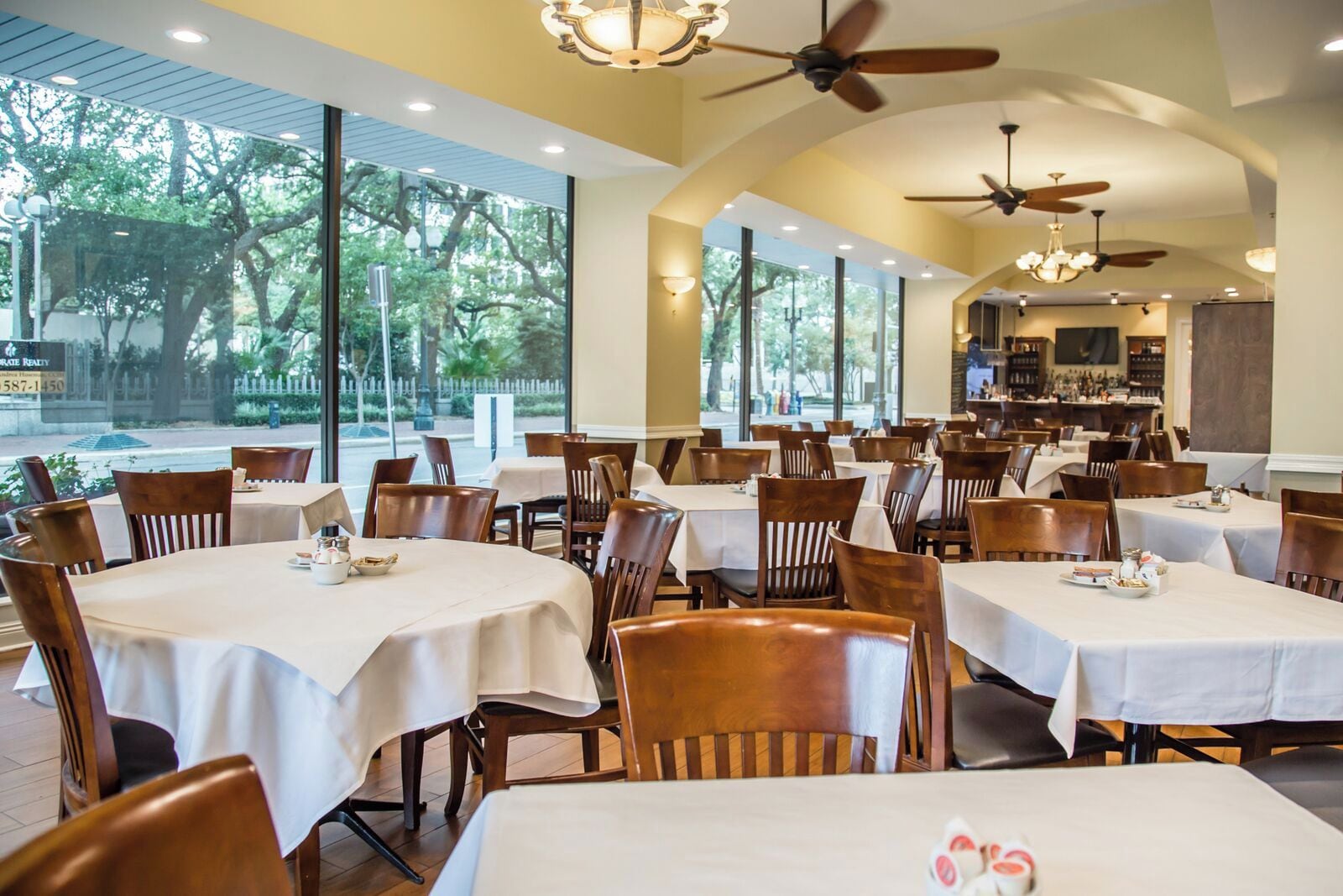 Restaurante com várias mesas e cadeiras, grandes janelas, ventiladores de teto e vista para o exterior.