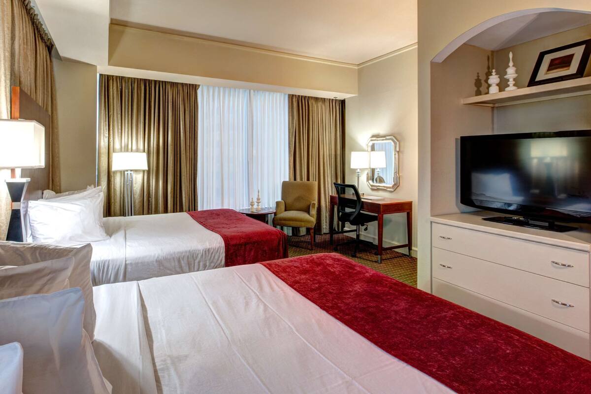 A hotel room with two beds, a desk, a chair, a television, and a minibar.