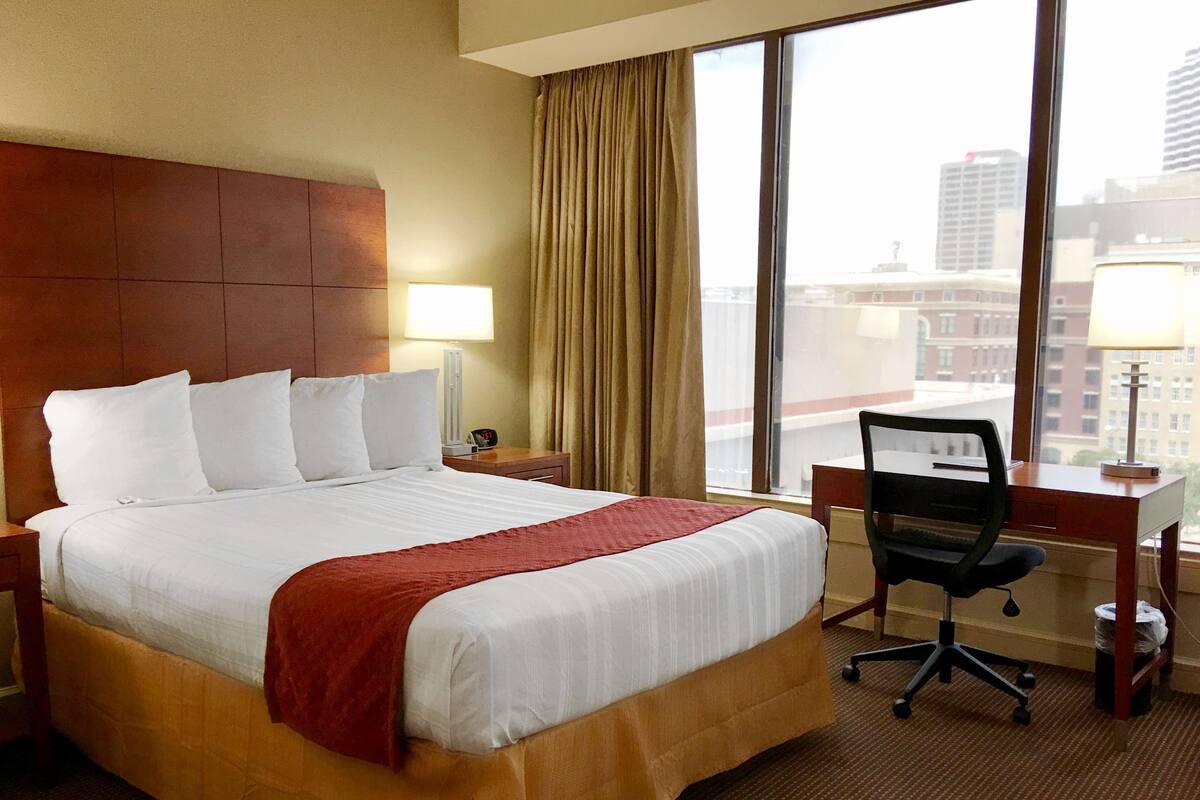 A hotel room with a large bed, a desk, a chair, and a view of the city through the window.