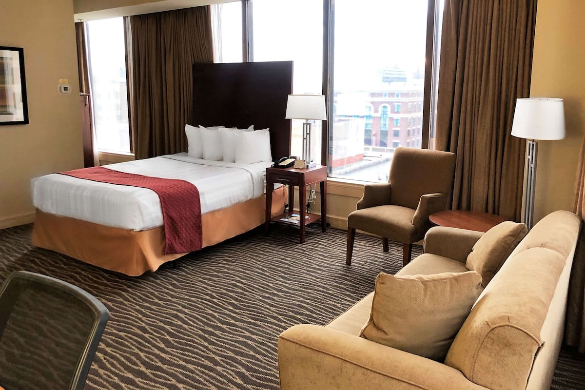 A hotel room with a large bed, a desk with a lamp, a chair, a sofa, and a view of a cityscape through large windows.