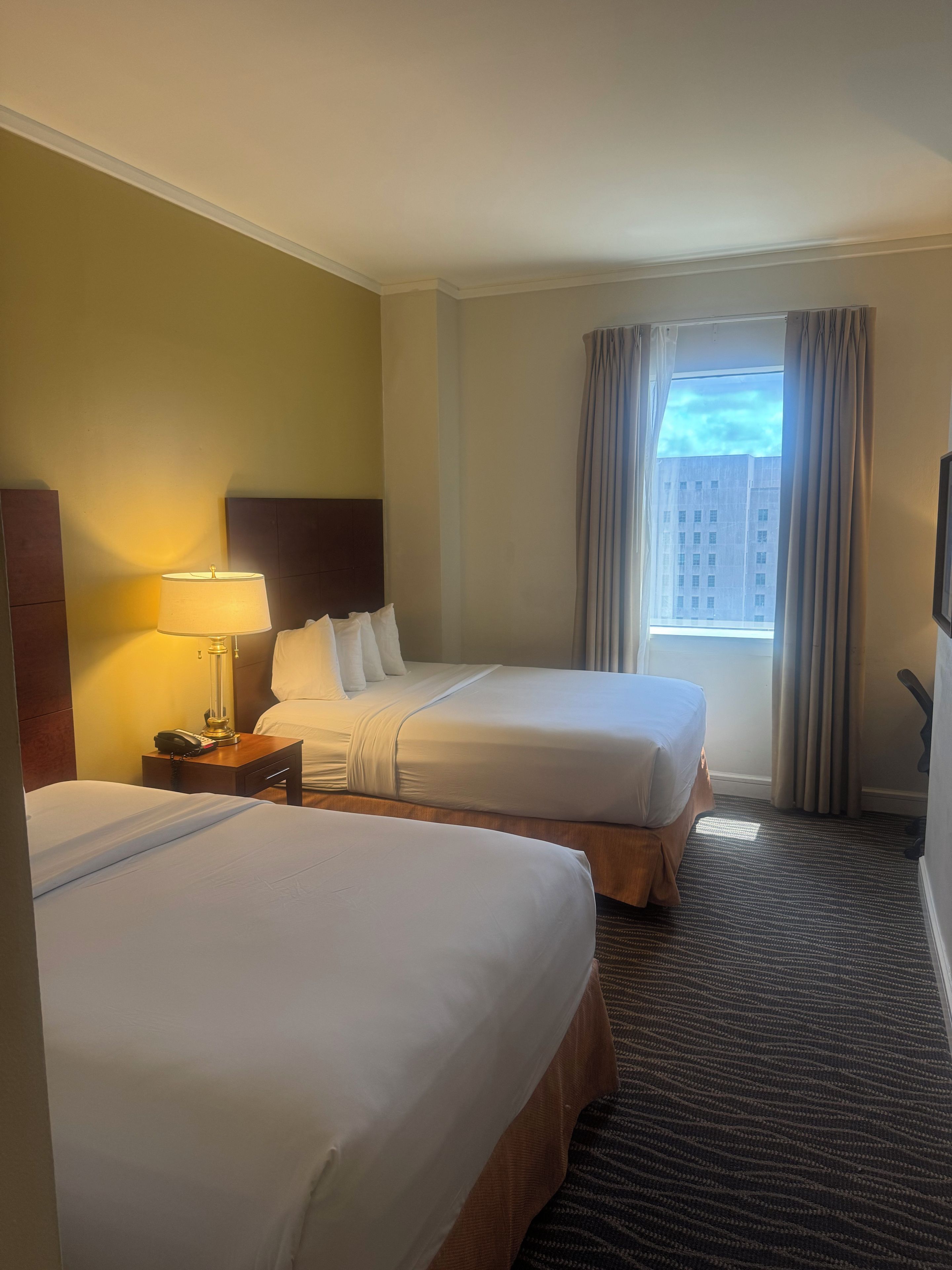 A hotel room with two beds, a nightstand with a lamp, a window with curtains, and a view of buildings outside.
