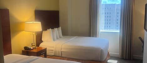 Une chambre d’hôtel avec deux lits, une table de chevet avec une lampe, une fenêtre avec des rideaux et une vue sur les bâtiments environnants.