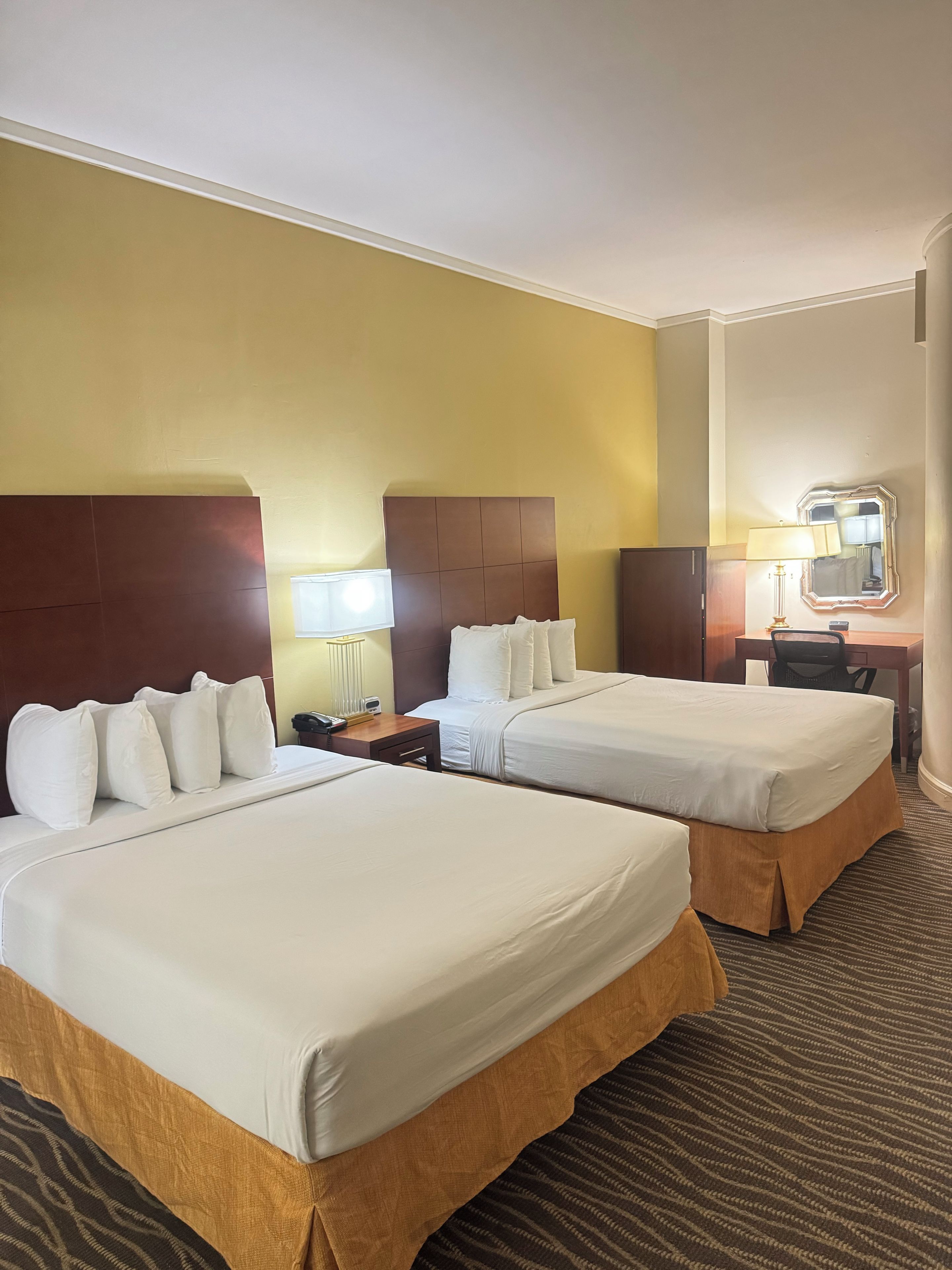 A hotel room with two beds, a desk, a chair, a lamp, and a mirror.