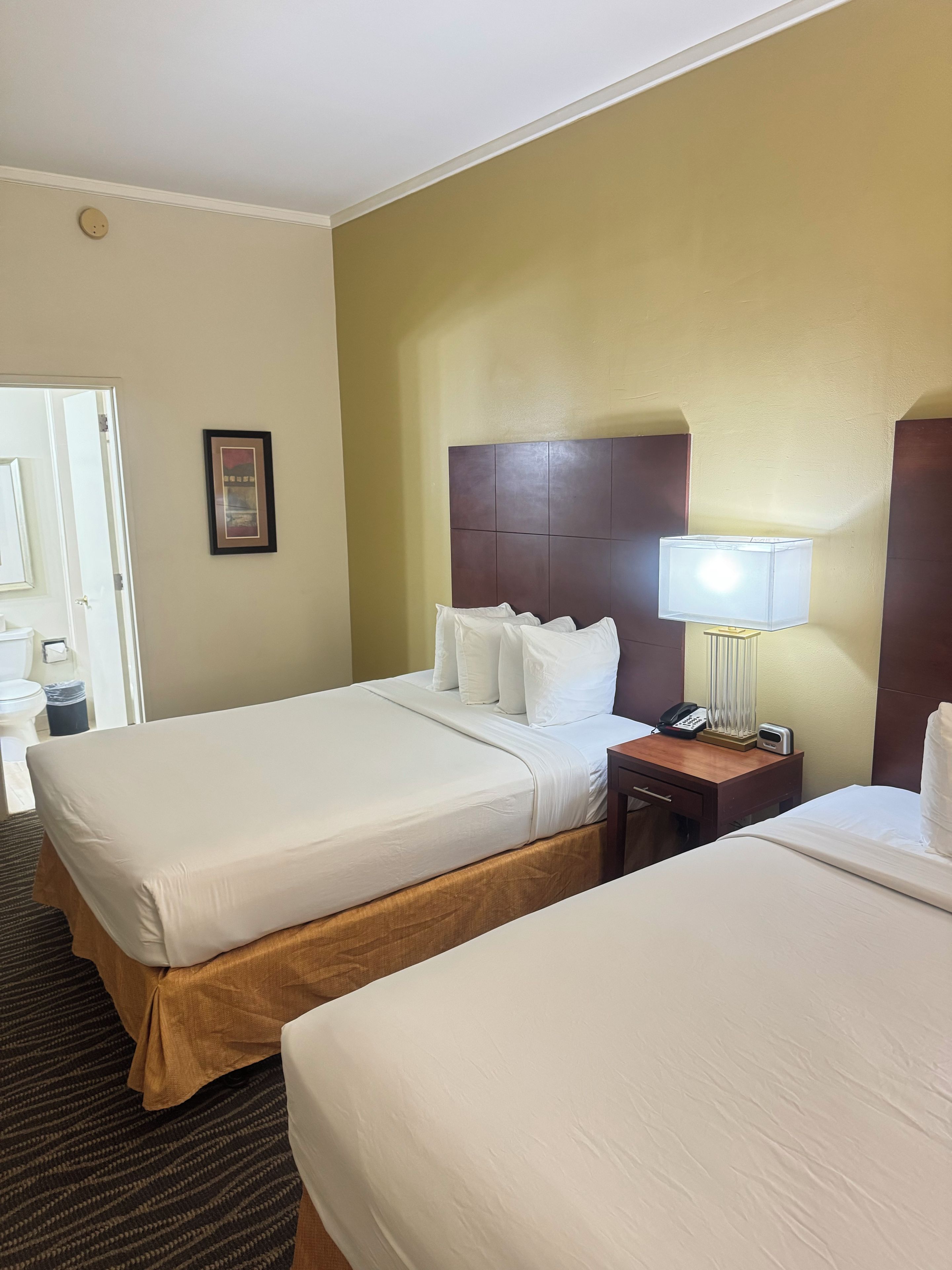 A hotel room with two beds, a bedside table with a lamp, a framed picture on the wall, and a door leading to the bathroom.
