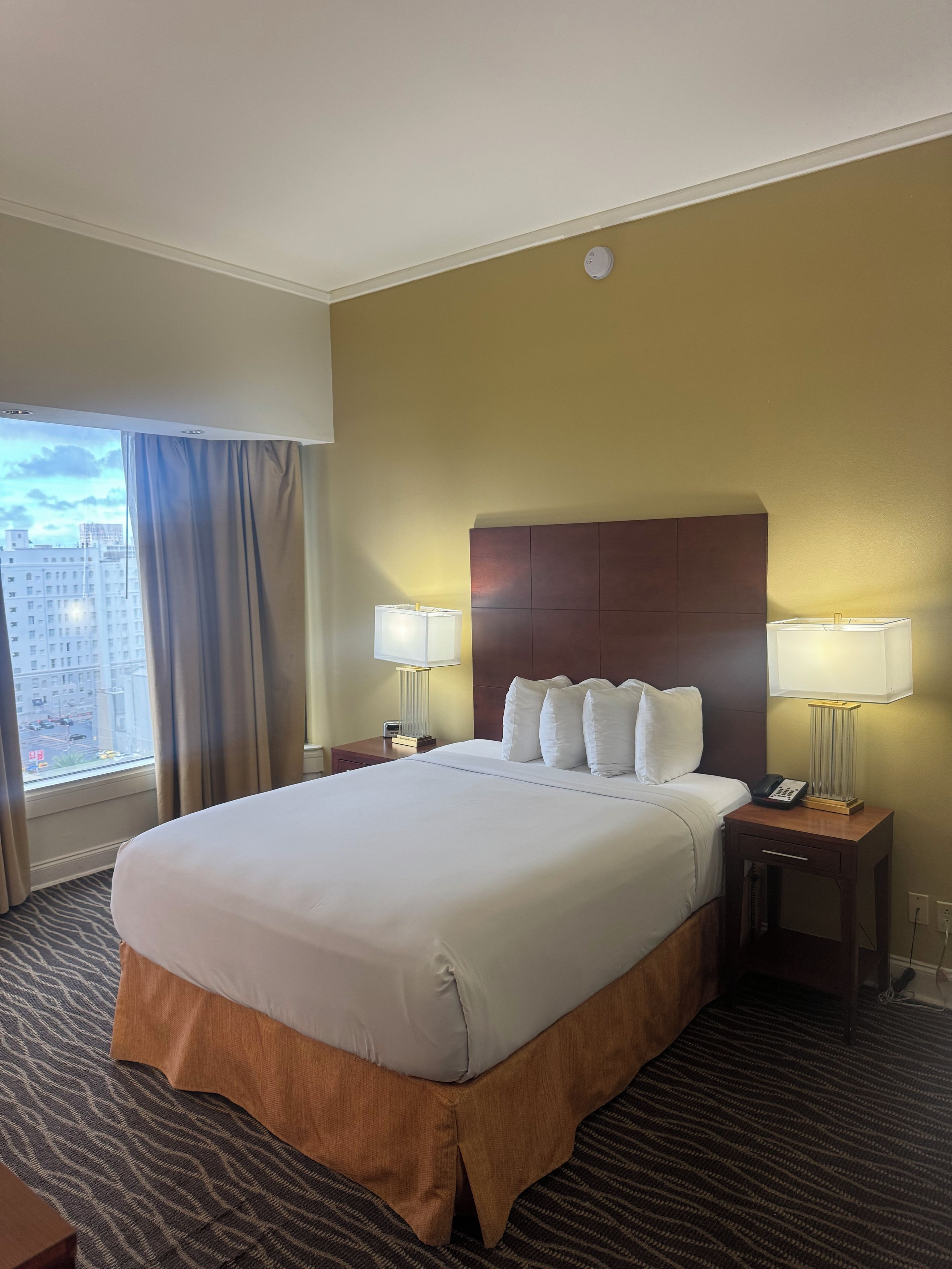 Habitación de hotel con una cama grande, dos mesitas de noche con lámparas, un cabecero y vistas a edificios a través de una ventana.