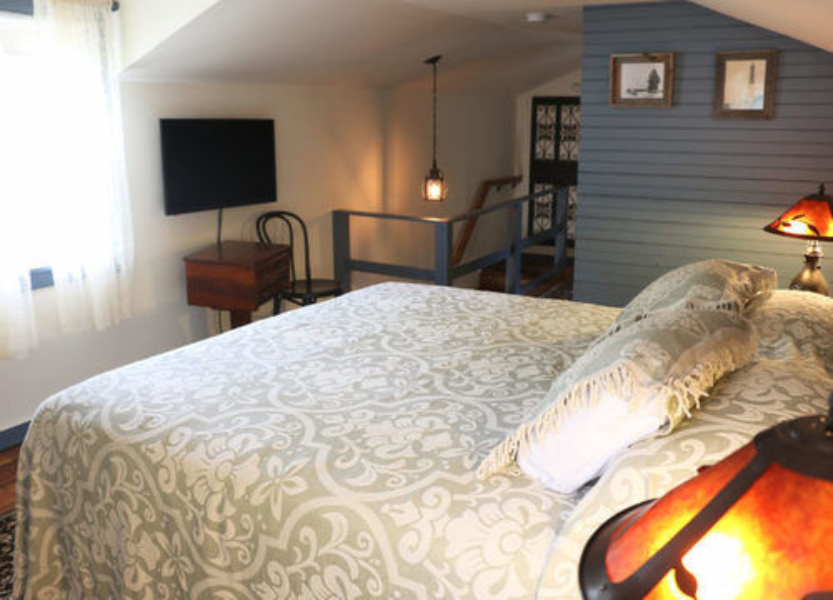 Une chambre avec une couvre-lit à motifs, une table de chevet avec une lampe, un téléviseur fixé au mur et un escalier menant à l’étage supérieur.