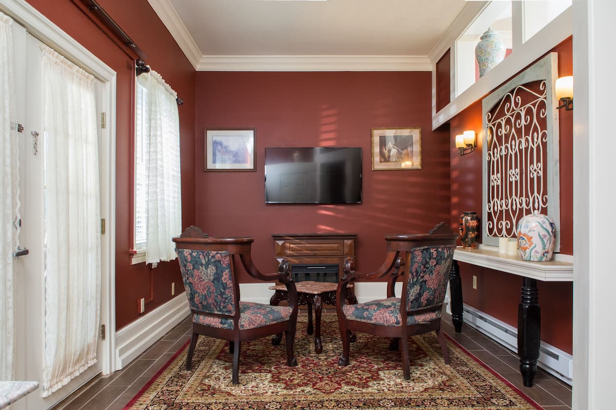 A room with a red wall, a flat-screen TV mounted above a wooden table with two chairs, and a patterned rug on the floor.