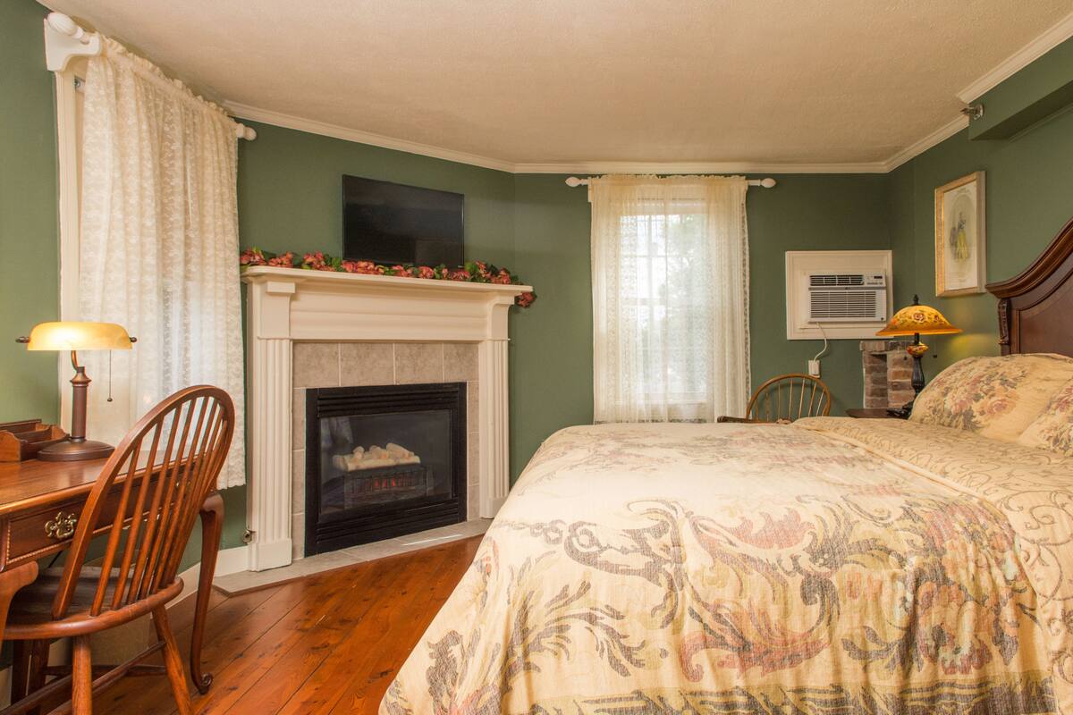 A room with a fireplace, a bed with a floral bedspread, a wooden chair, and a desk with a lamp.