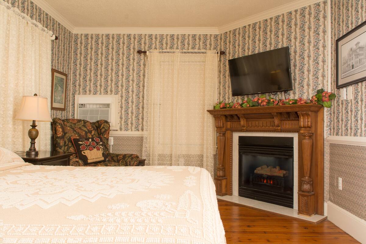 A bedroom with a large bed, a fireplace, a television mounted above it, and floral wallpaper.