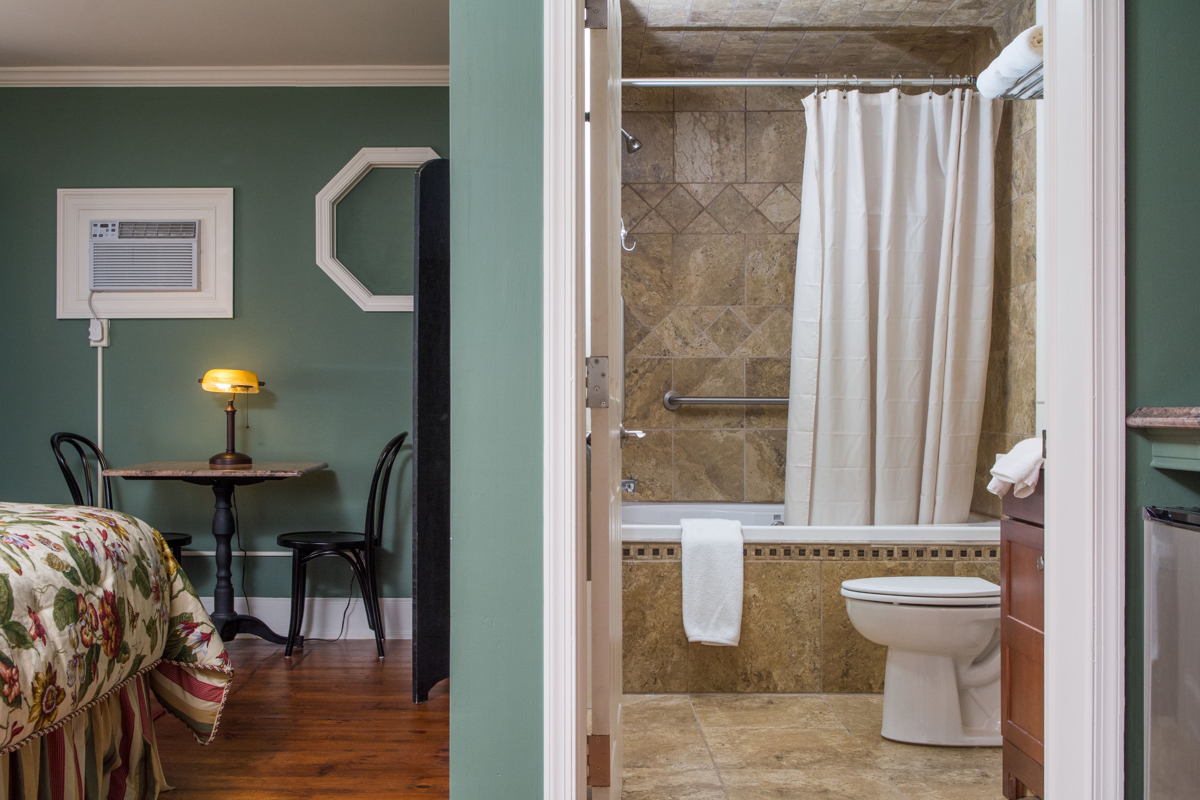 A room with a bed, a table with a lamp, a shower area with a bathtub, and a toilet.