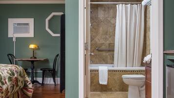 A room with a bed, a table with a lamp, a shower area with a bathtub, and a toilet.