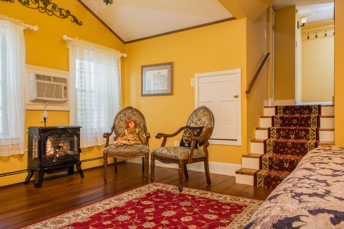 A room with yellow walls, a red patterned rug, two wooden chairs, a vintage stove, and a staircase with red carpet.