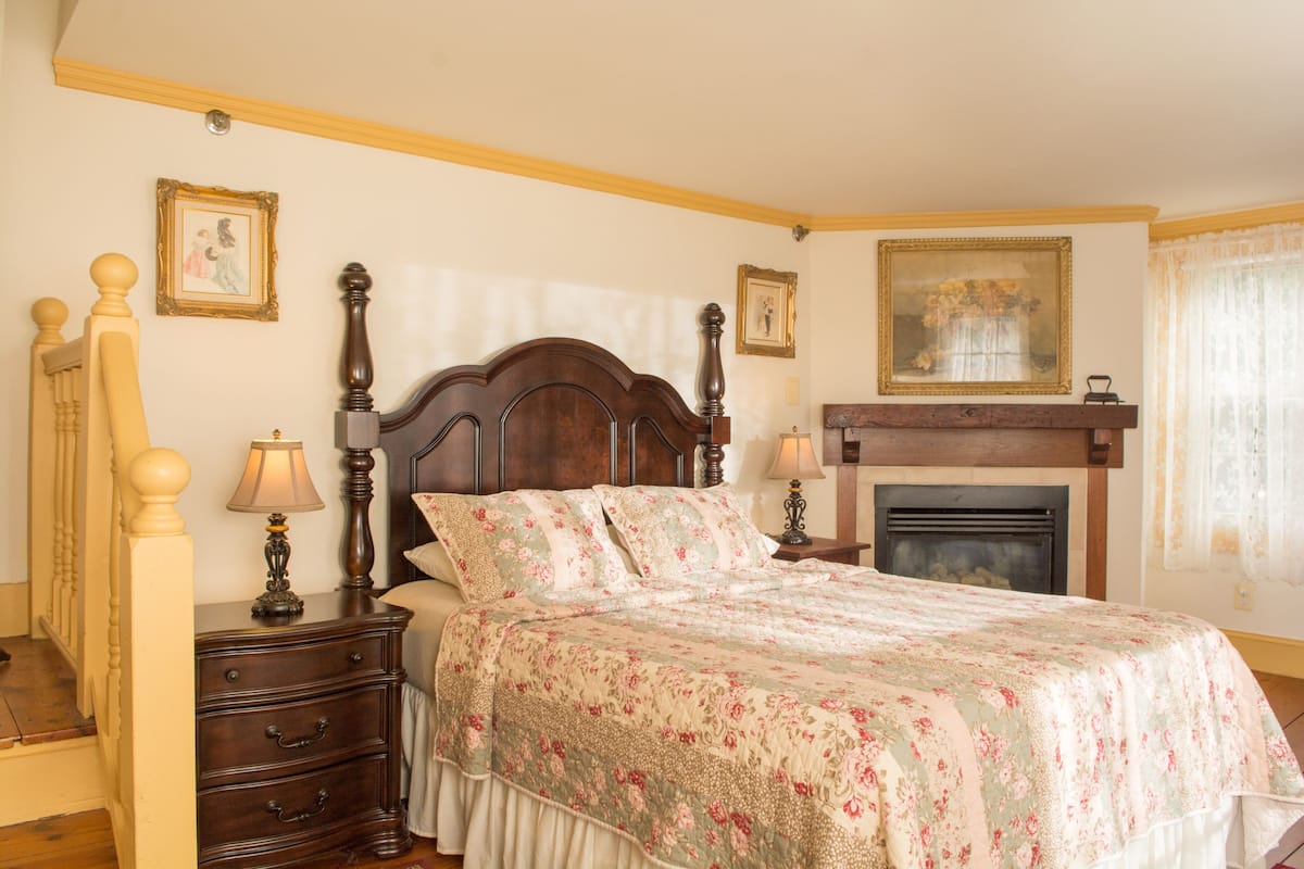 A bedroom with a large bed, wooden headboard, two bedside tables with lamps, a fireplace, and framed pictures on the walls.