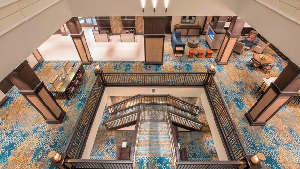 Eine geräumige Lobby mit einer zentralen Treppe, Sitzbereichen und einem gemusterten Teppich.