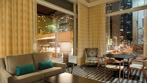 Ein Hotelzimmer mit einer Couch, Sessel, einem Tisch und Blick auf die beleuchtete Stadt bei Nacht.