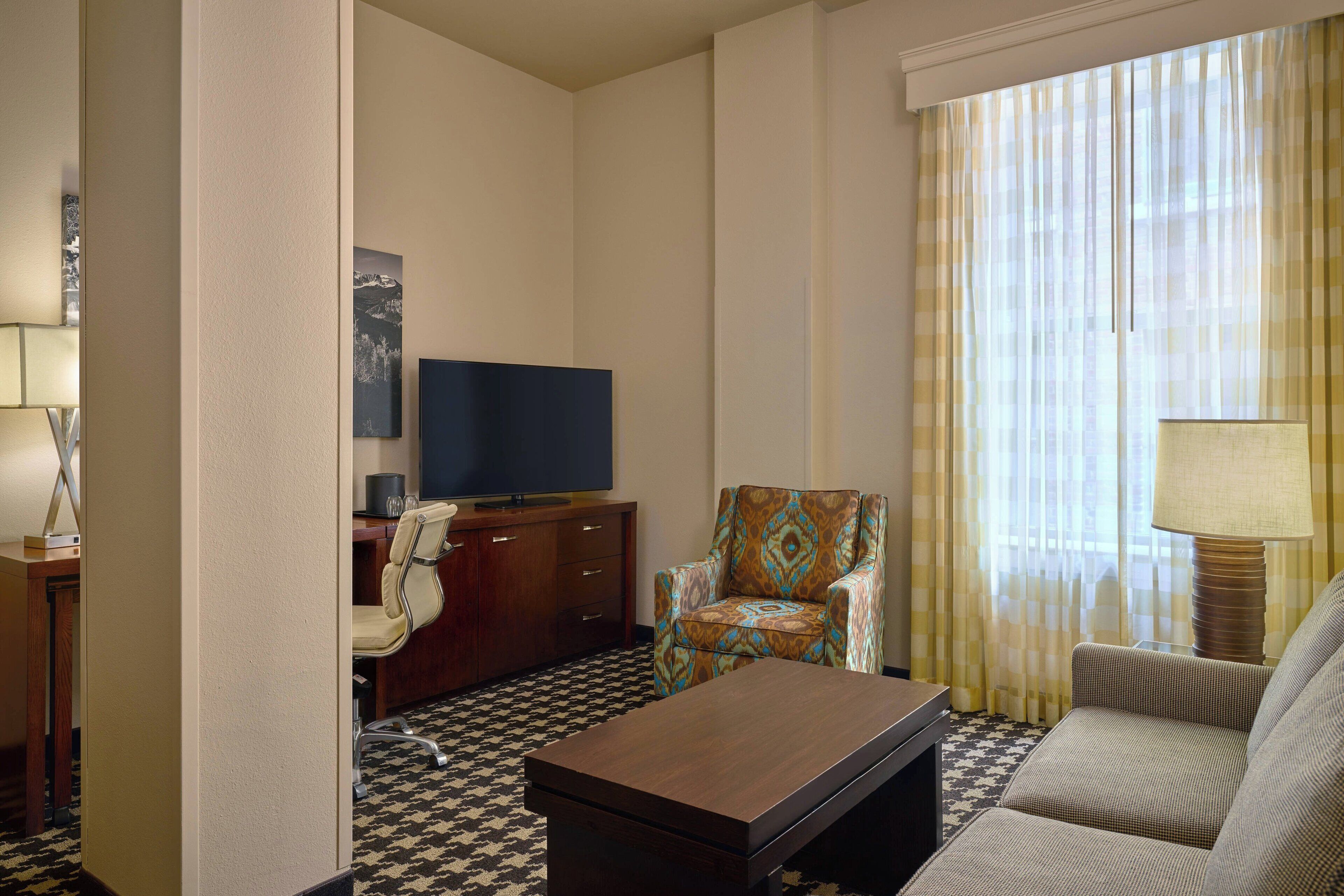 Habitación de hotel con suelo de cuadros, televisor de pantalla plana, sillón estampado, sofá y lámpara.