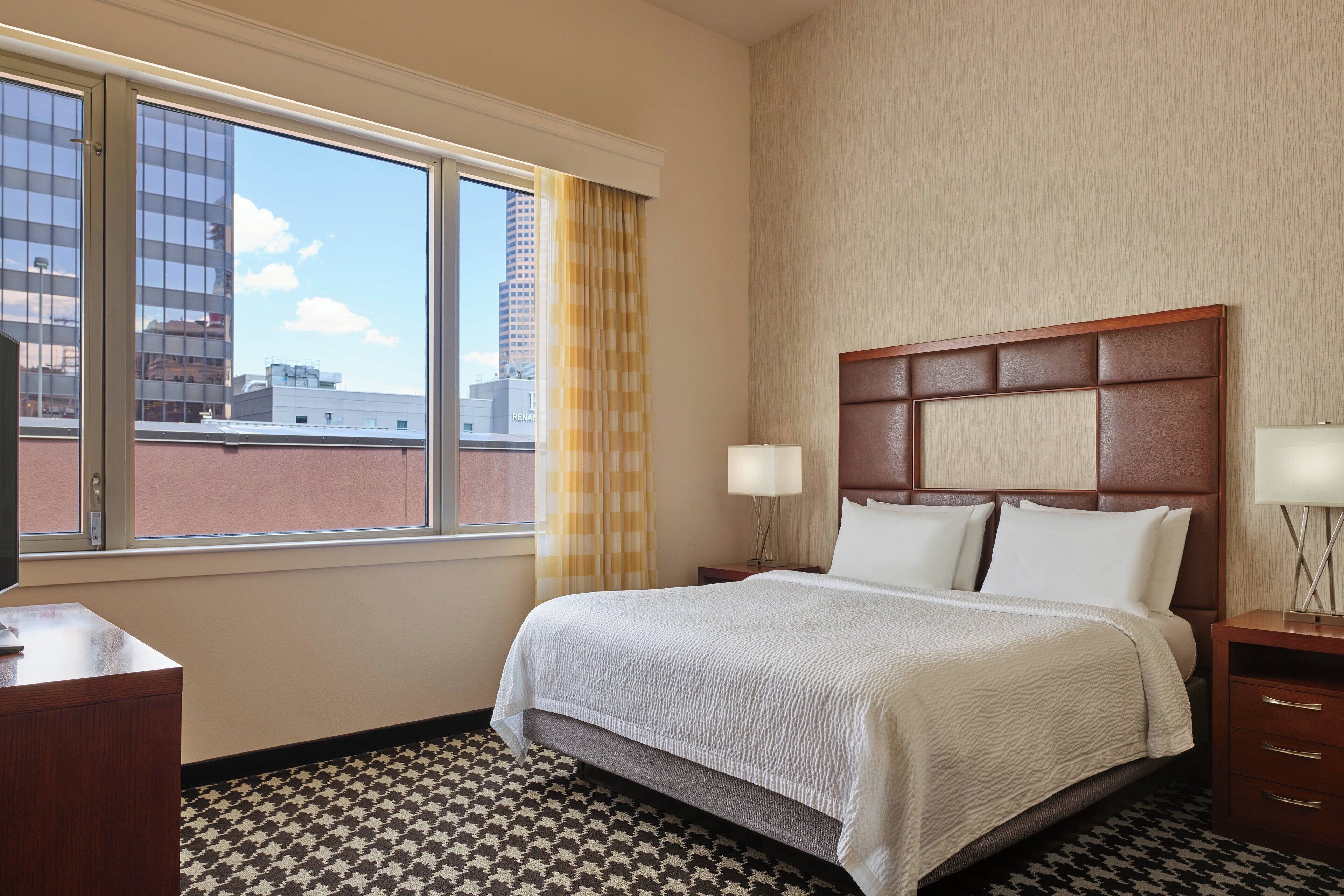 A hotel room with a large bed, a nightstand, a desk, and a window with a city view.