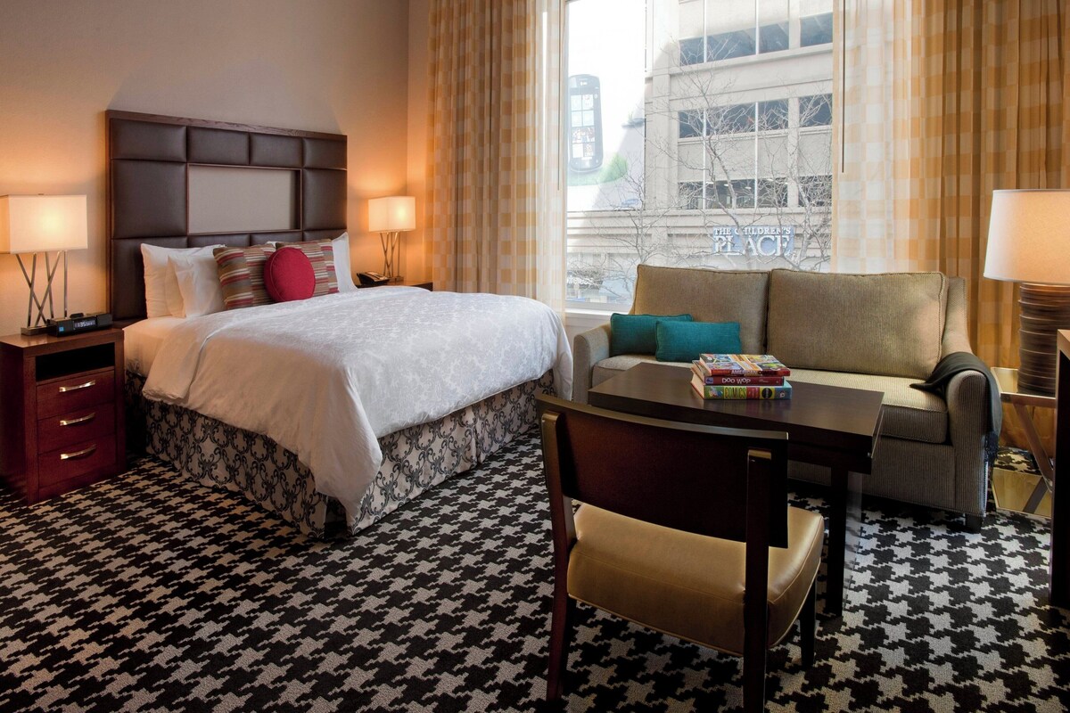 A hotel room with a large bed, a sofa, a small table, and a chair. There is a window with curtains and a view of buildings outside.
