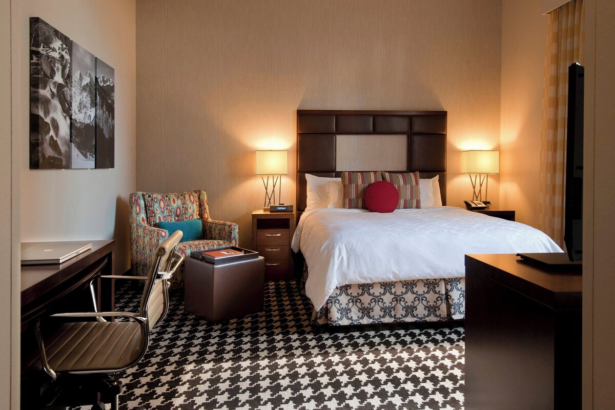 A hotel room with a bed, a desk, a chair, a TV, and a lamp.