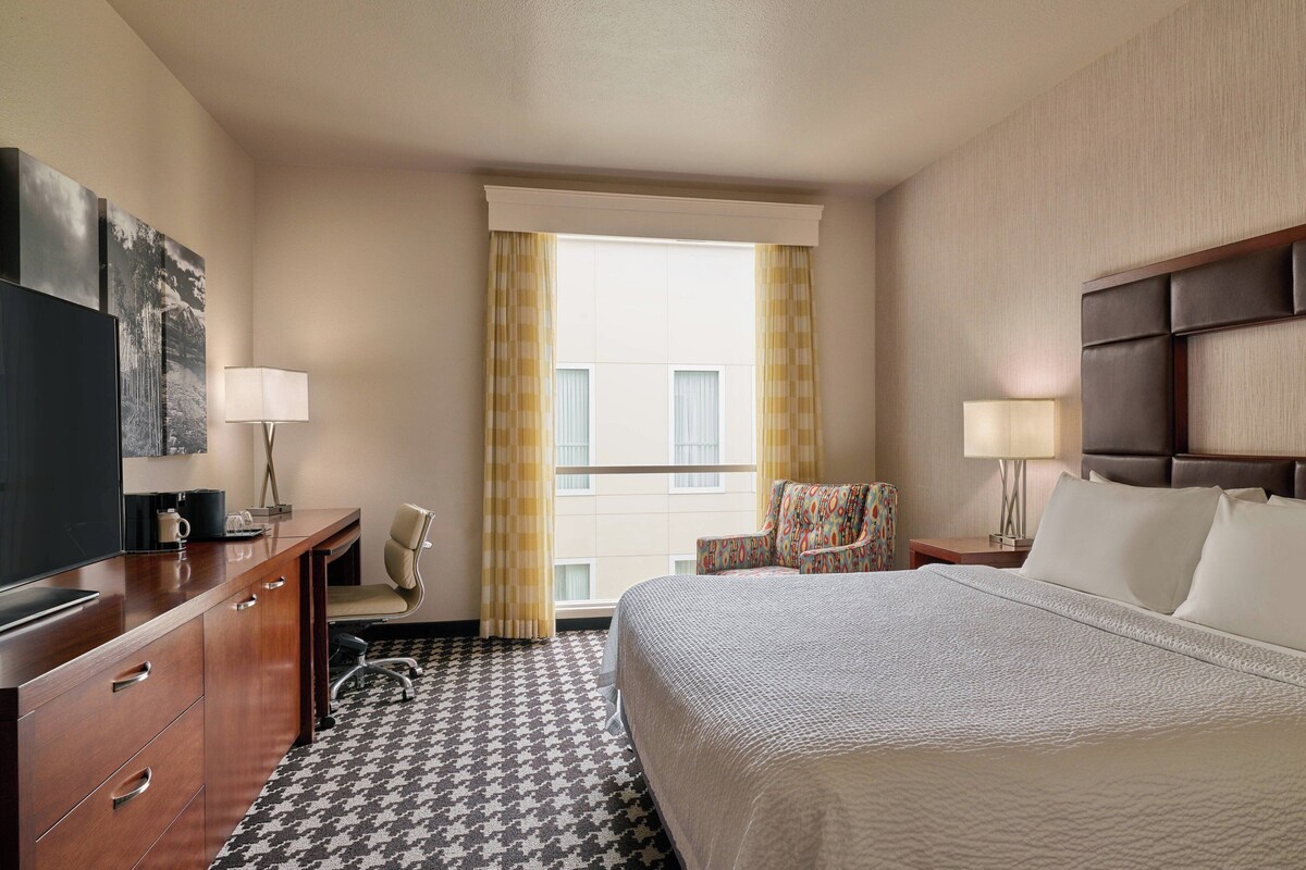 A hotel room with a large bed, a desk with a television, a chair, and a window with curtains.