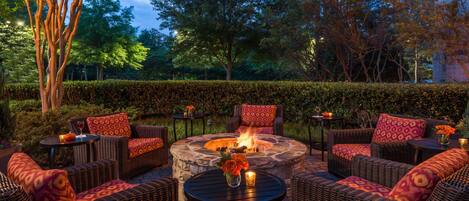 An outdoor seating area with wicker furniture, a stone fire pit, and a hedge in the background.