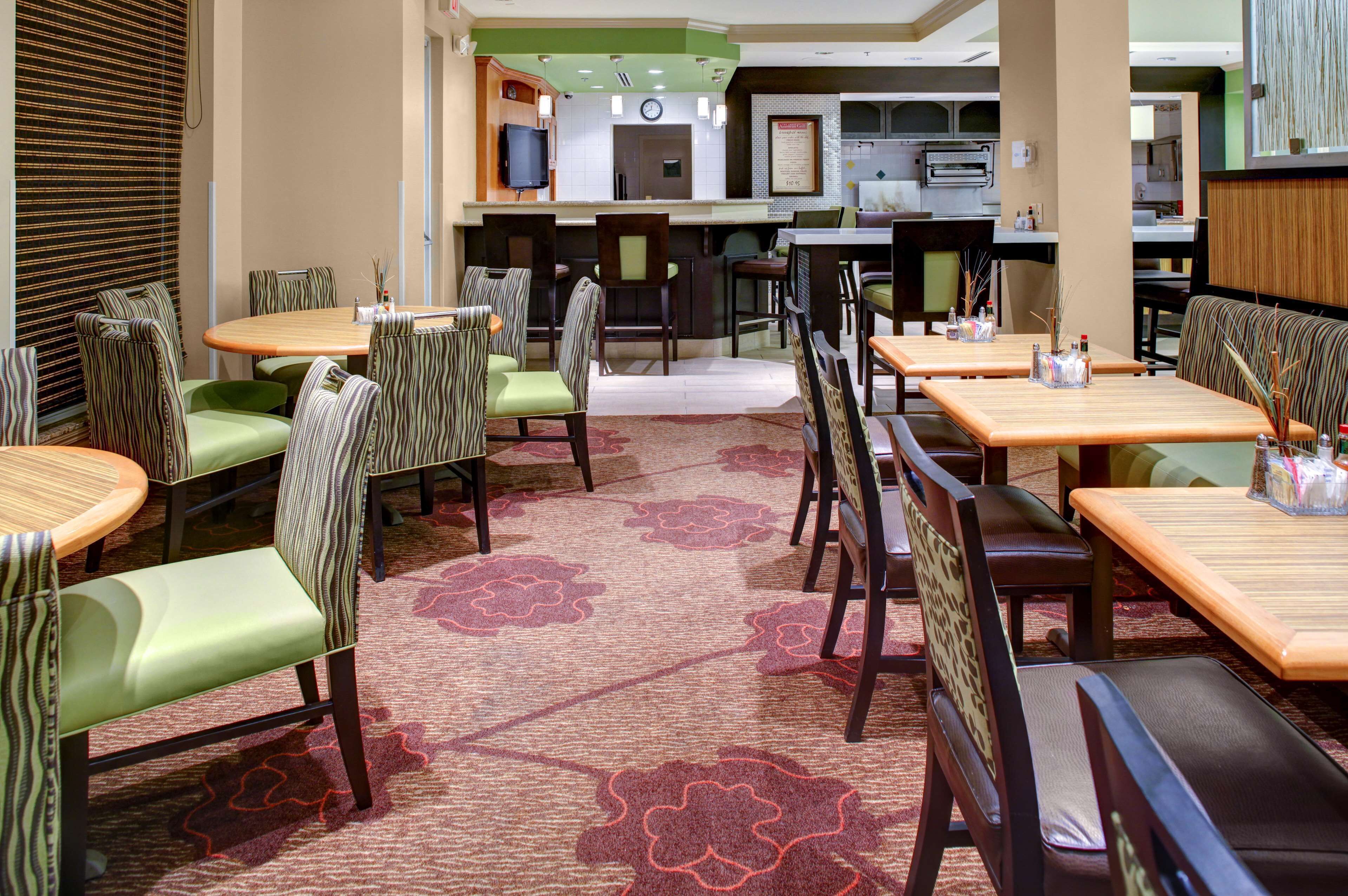 A restaurant with multiple dining tables and chairs, a kitchen area in the background, and a patterned carpet.
