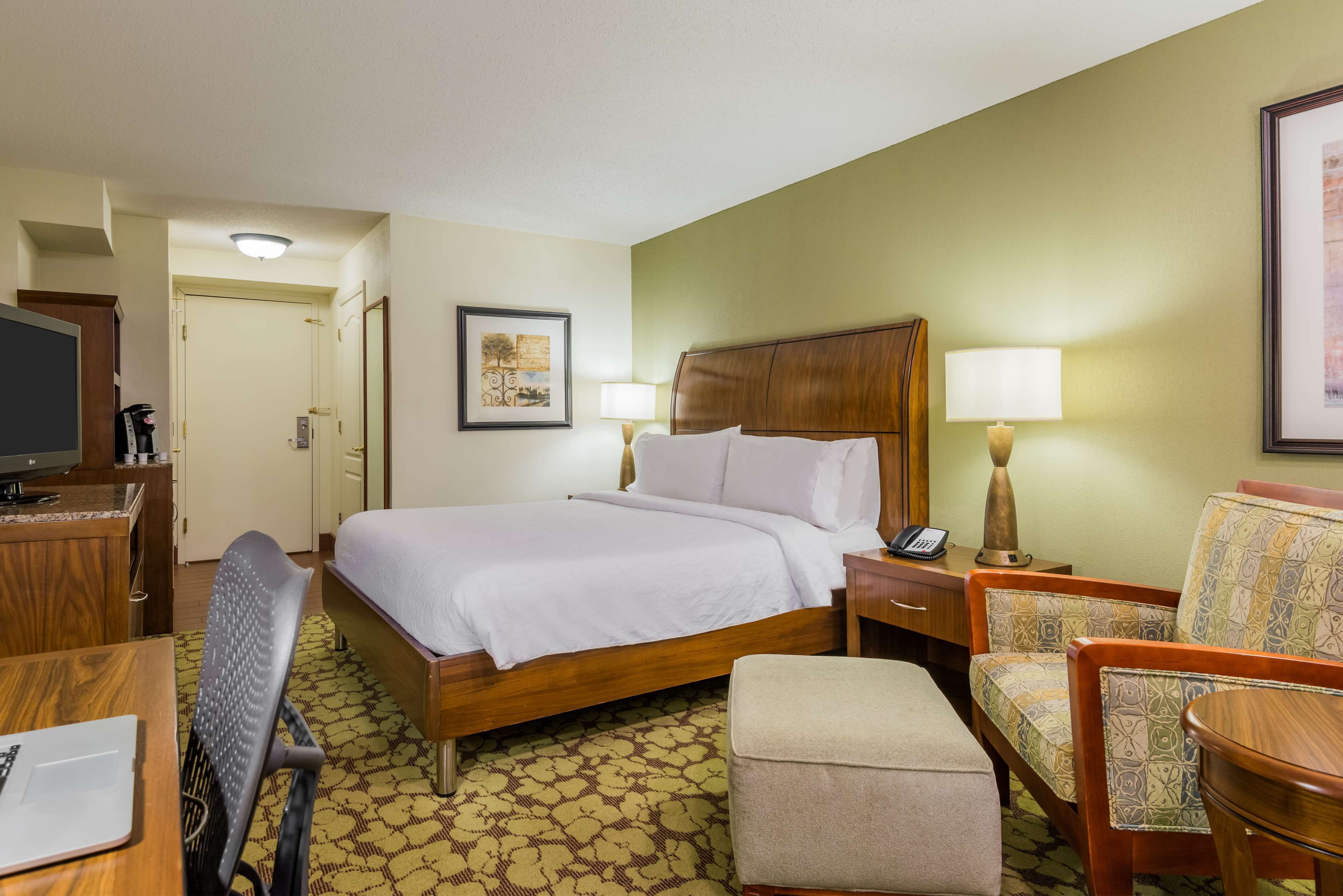 A hotel room with a bed, a desk with a computer, a chair, a small table, and a lamp.