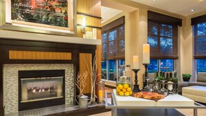 A modern living room with a fireplace, a coffee table with drinks and snacks, and a large window with blinds. - Hilton Garden Inn Atlanta North/Alpharetta (Alpharetta)