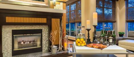 A modern living room with a fireplace, a coffee table with drinks and snacks, and a large window with blinds.