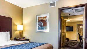 Une chambre d’hôtel comprenant un lit, une lampe de chevet, un téléphone, un tableau encadré au mur, et une porte donnant sur un couloir où se trouvent un téléviseur et un bureau.
