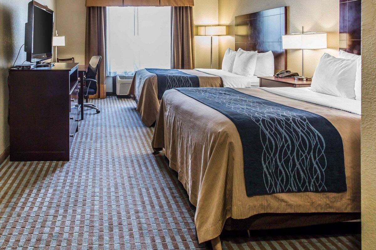 A hotel room with two beds, a desk with a computer, a TV, and a large window with curtains.