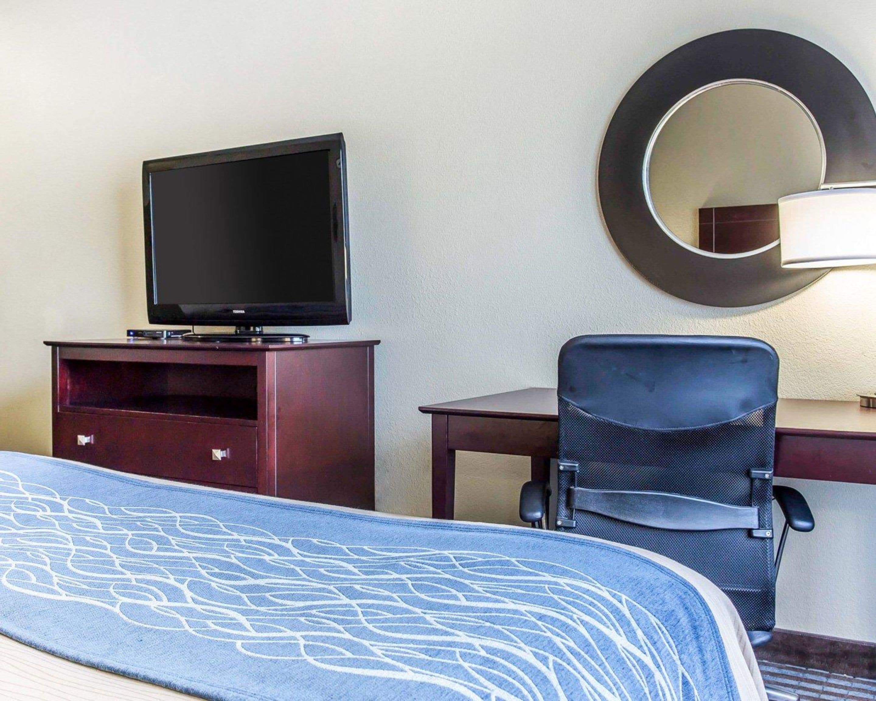 A hotel room with a bed, a TV on a wooden cabinet, a desk with a chair, and a wall-mounted mirror.