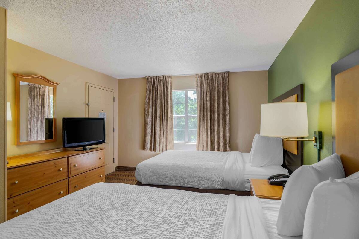Habitación de hotel con dos camas, televisor, escritorio con lámpara y ventana con cortinas.