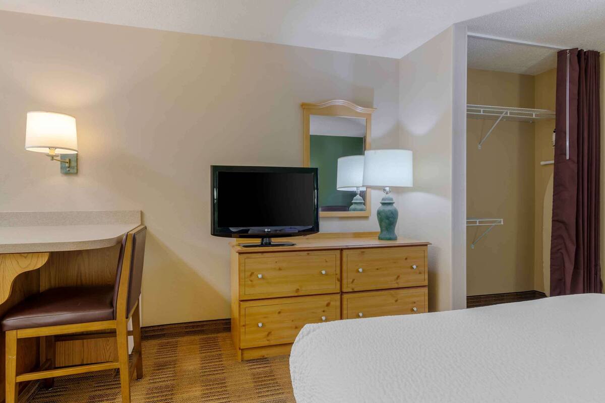 Una habitación de hotel con cama, escritorio y silla, televisor, cómoda, espejo y zona de ducha.