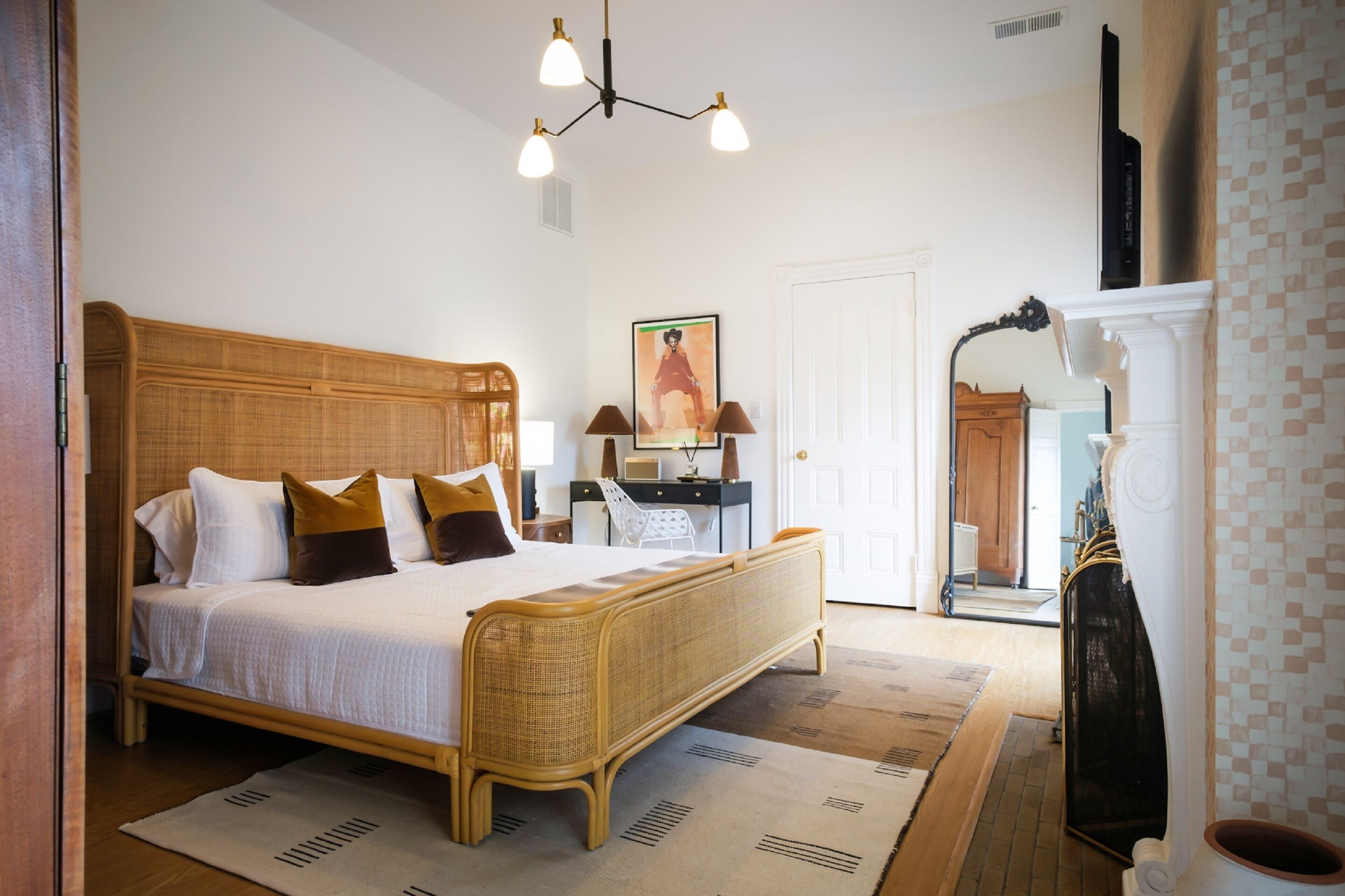 A bedroom with a wooden bed, a desk, and a chair. There is a painting on the wall and a door with a mirror.