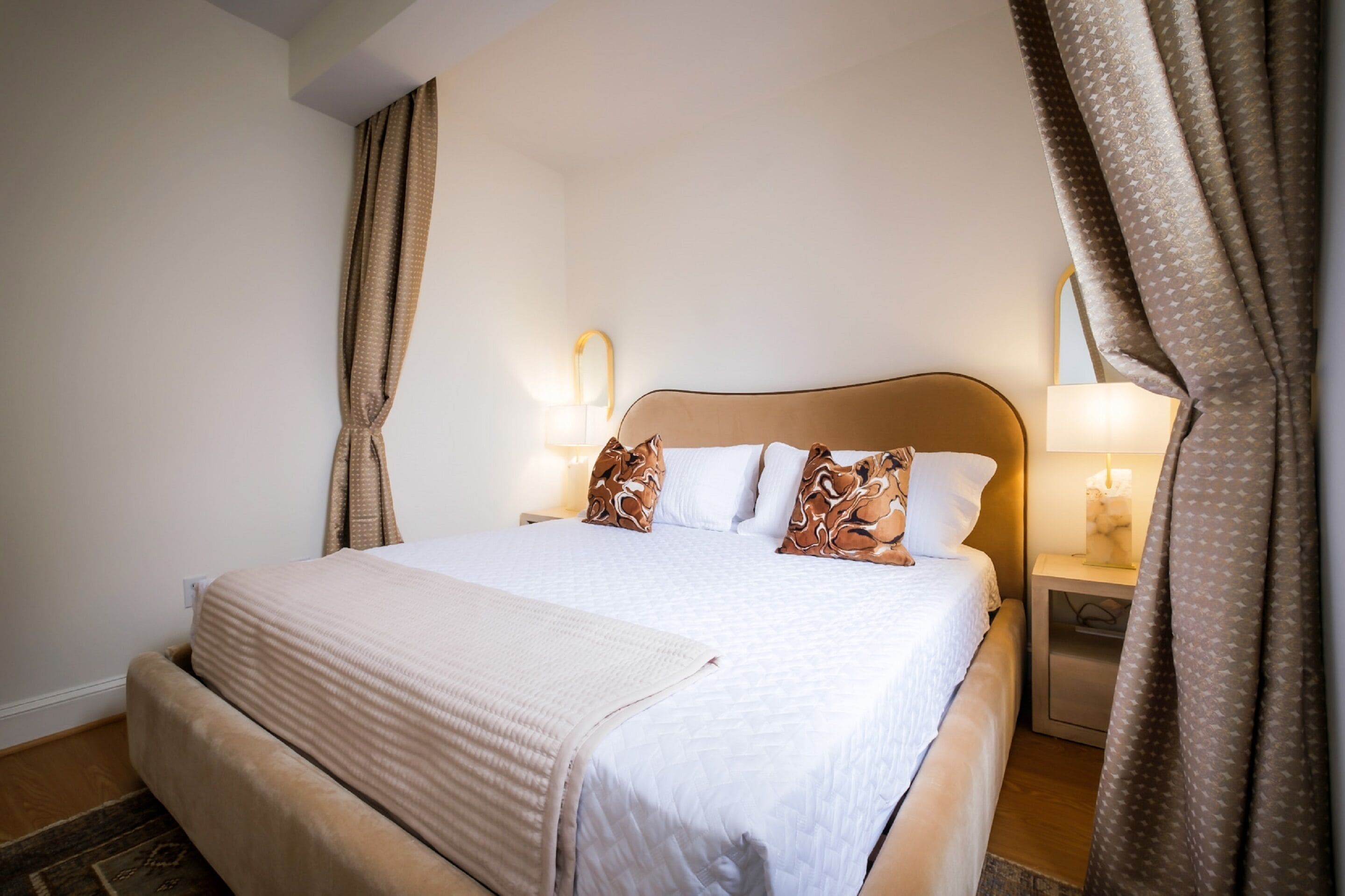A neatly made bed with a beige blanket, two bedside lamps, and a nightstand with a tissue box.