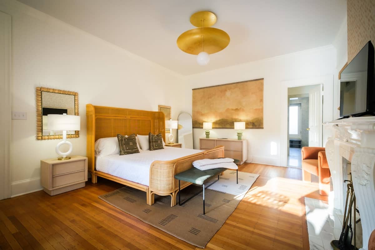 A spacious bedroom with a large bed, bedside tables, a bench, a fireplace, and a painting on the wall.