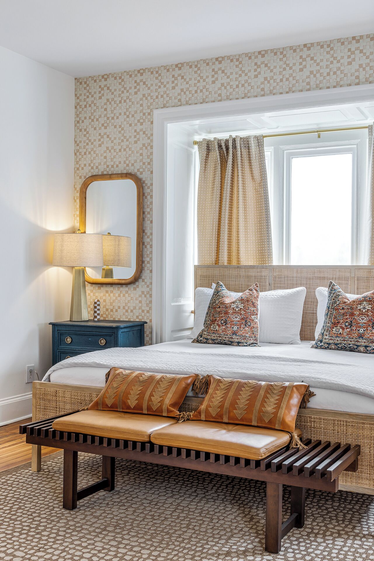 Een slaapkamer met een groot bed, een nachtkastje met lamp, een spiegel, behang met patroon, een raam met gordijnen en een bank met kussens.