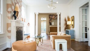 Ein geräumiges Wohnzimmer mit Kamin, einem runden Hocker, einer Couch und einem Couchtisch. Darüber hängt eine Kristallleuchter und an der Wand befindet sich ein großer Spiegel.