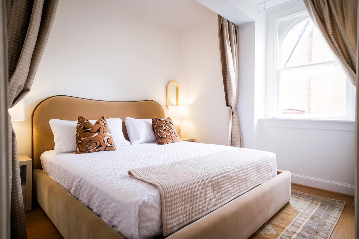 A neatly made bed with a headboard, a bedside lamp, and a window with curtains.