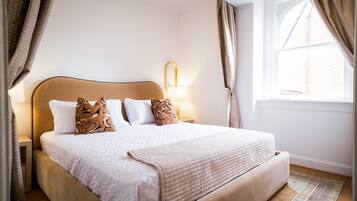 A neatly made bed with a headboard, a bedside lamp, and a window with curtains.