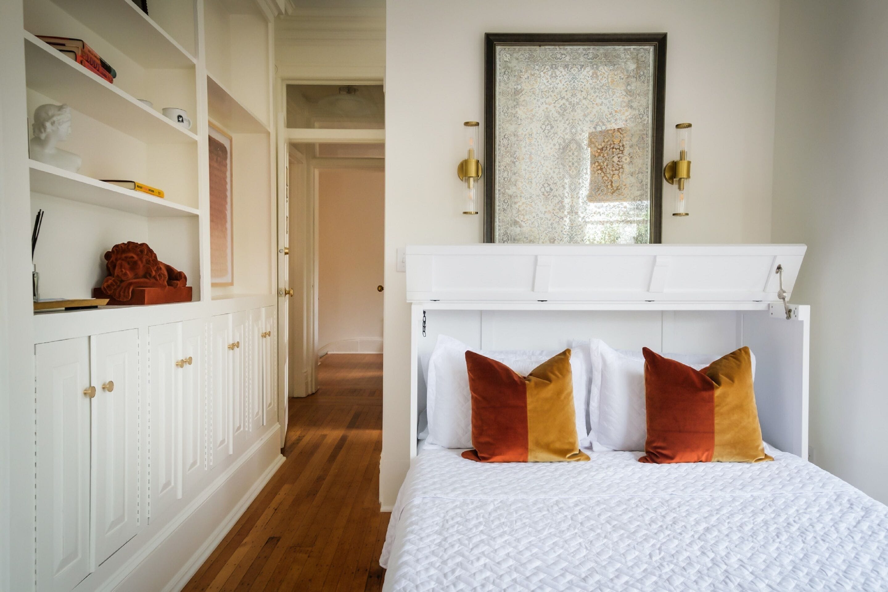 Une chambre à coucher avec un lit blanc, deux oreillers, une armoire blanche et un miroir doté d’un cadre à motifs.
