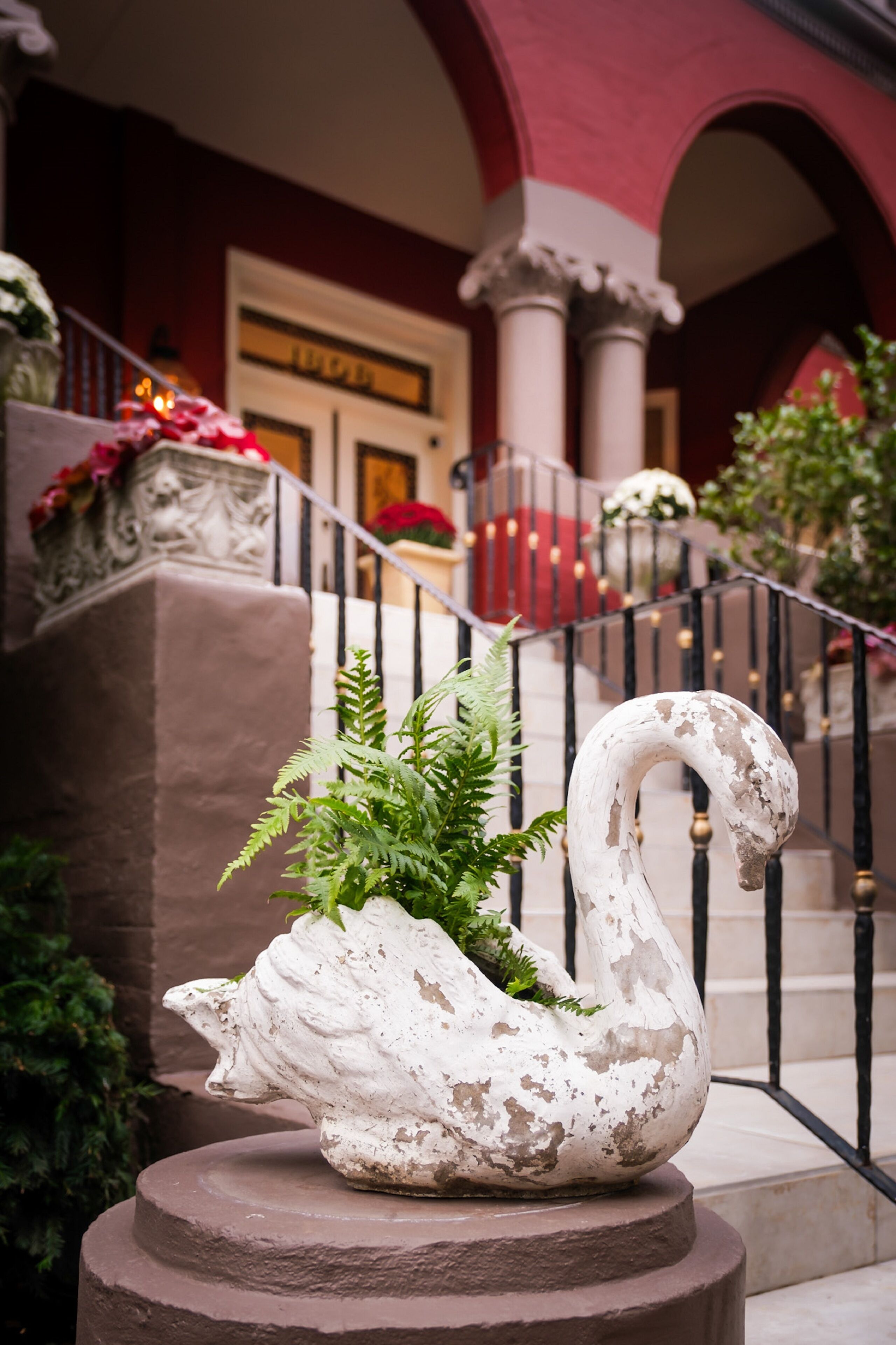 A swan-shaped planter with greenery on a pedestal in a courtyard with a staircase and red walls.