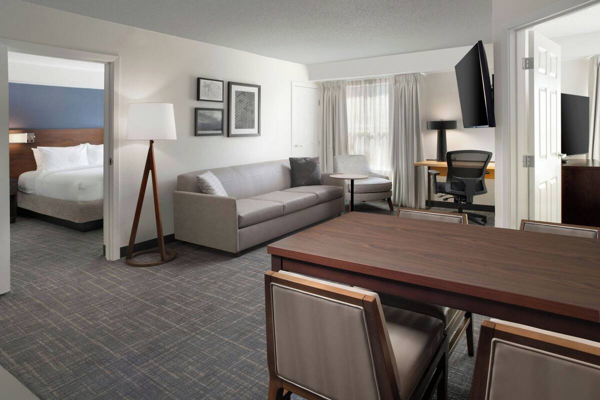 Ein Hotelzimmer mit Bett, Sofa, Esstisch mit Stühlen, Fernseher und Schreibtisch mit Sessel.