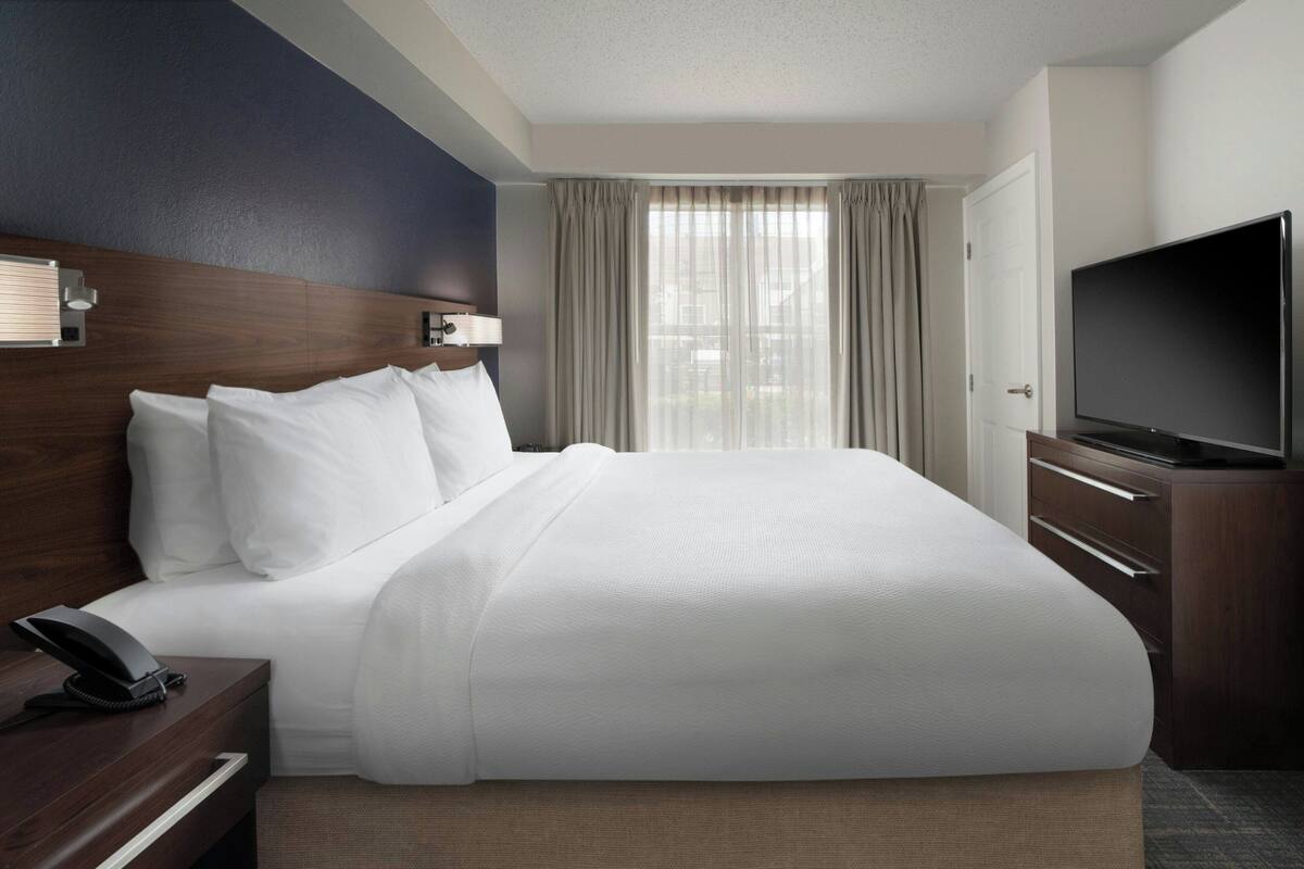 Ein Hotelzimmer mit einem großen Bett, einem Fernseher, einem Schreibtisch mit Telefon und einem Fenster mit Vorhängen.