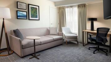 Quarto de hotel com sofá, poltrona, escrivaninha e televisão de tela plana.