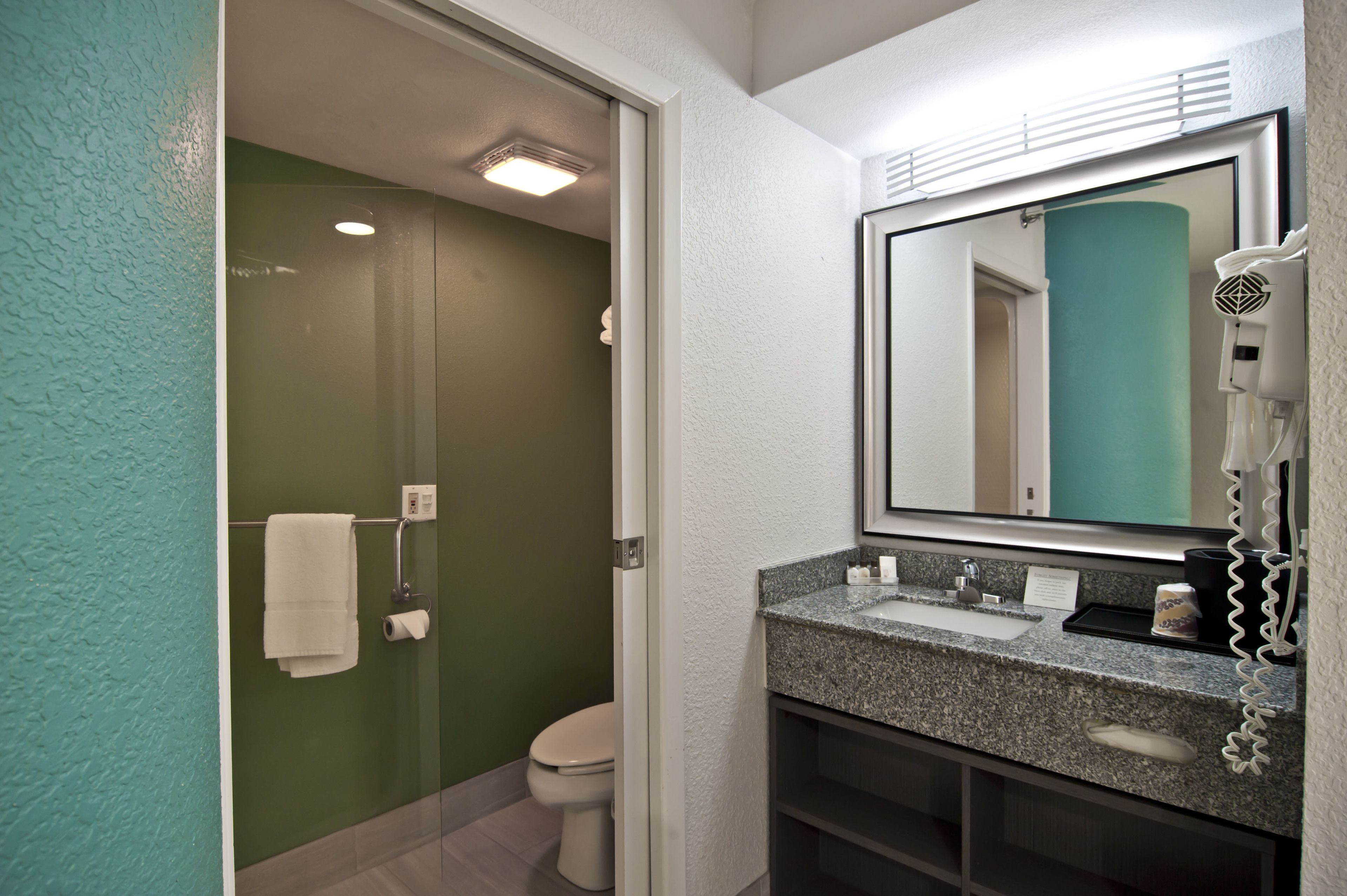 A bathroom with a glass shower enclosure, a white toilet, a granite countertop sink, a hairdryer, and a mirror.